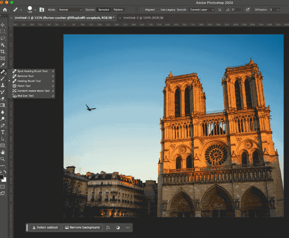 Using the healing brush tool in Photoshop to remove a bird from the sky in a photo of a tall building with a clear blue sky with one bird in the sky 