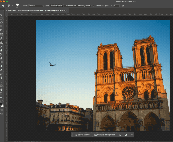 Using the spot healing brush tool in photoshop to remove a bird from the sky in a photo of a tall building with a clear blue sky with one bird in the sky 